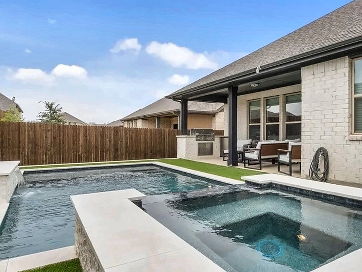 Pool with a raised spa, stacked-stone wall and artificial turf beside a covered patio with an outdoor kitchen